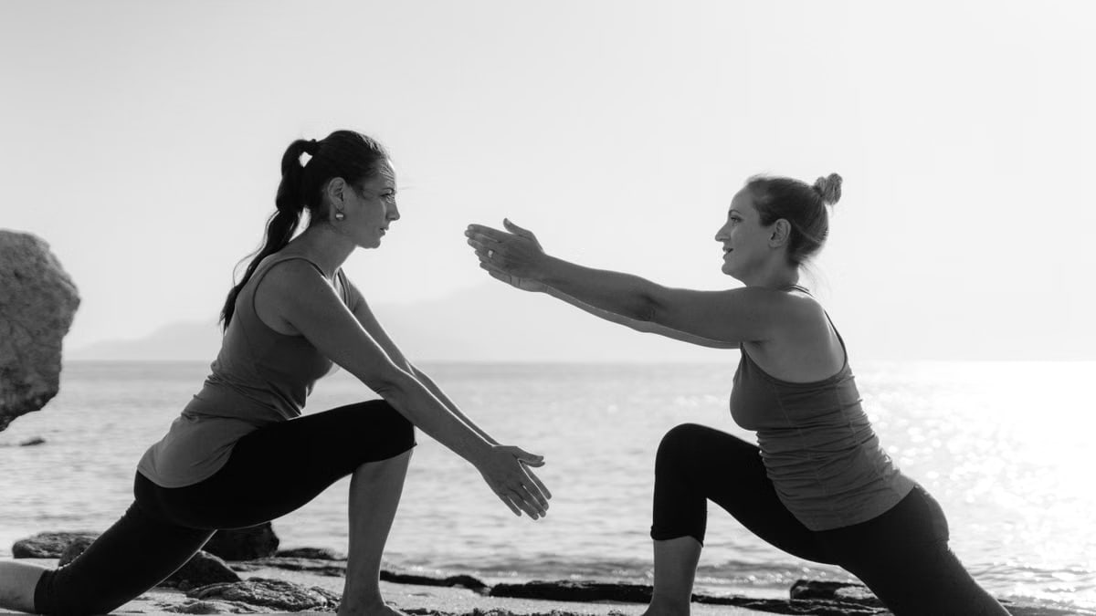 Two women performing yoga on a sandy beach during sunrise, promoting wellness and fitness.